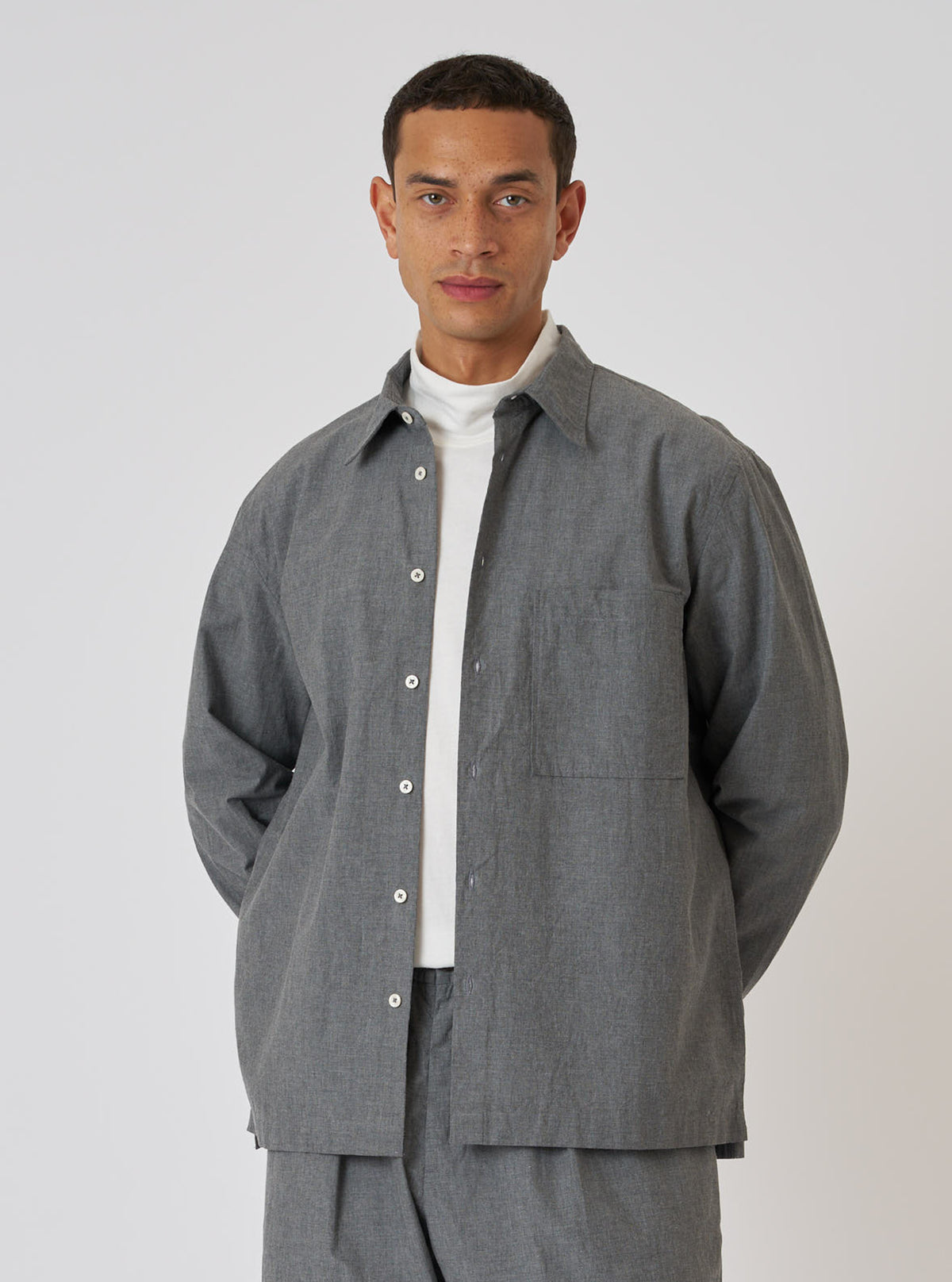 A model poses in a Charcoal Organic Chambray Lucky Shirt and matching pants against a white backdrop. The shirt is a relaxed fit with a button-down front.