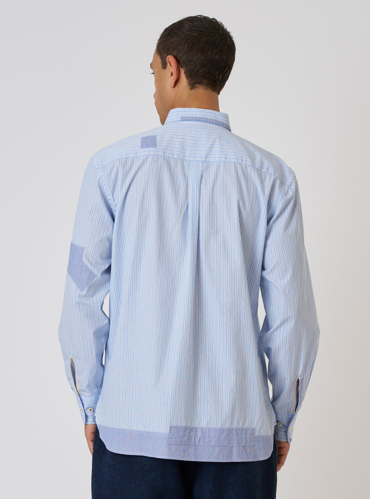 Back view of a model wearing the Blue Office Stripe Patched Lazy Day Shirt. The shirt is blue and white striped with patched detailing. Shot against a white background.