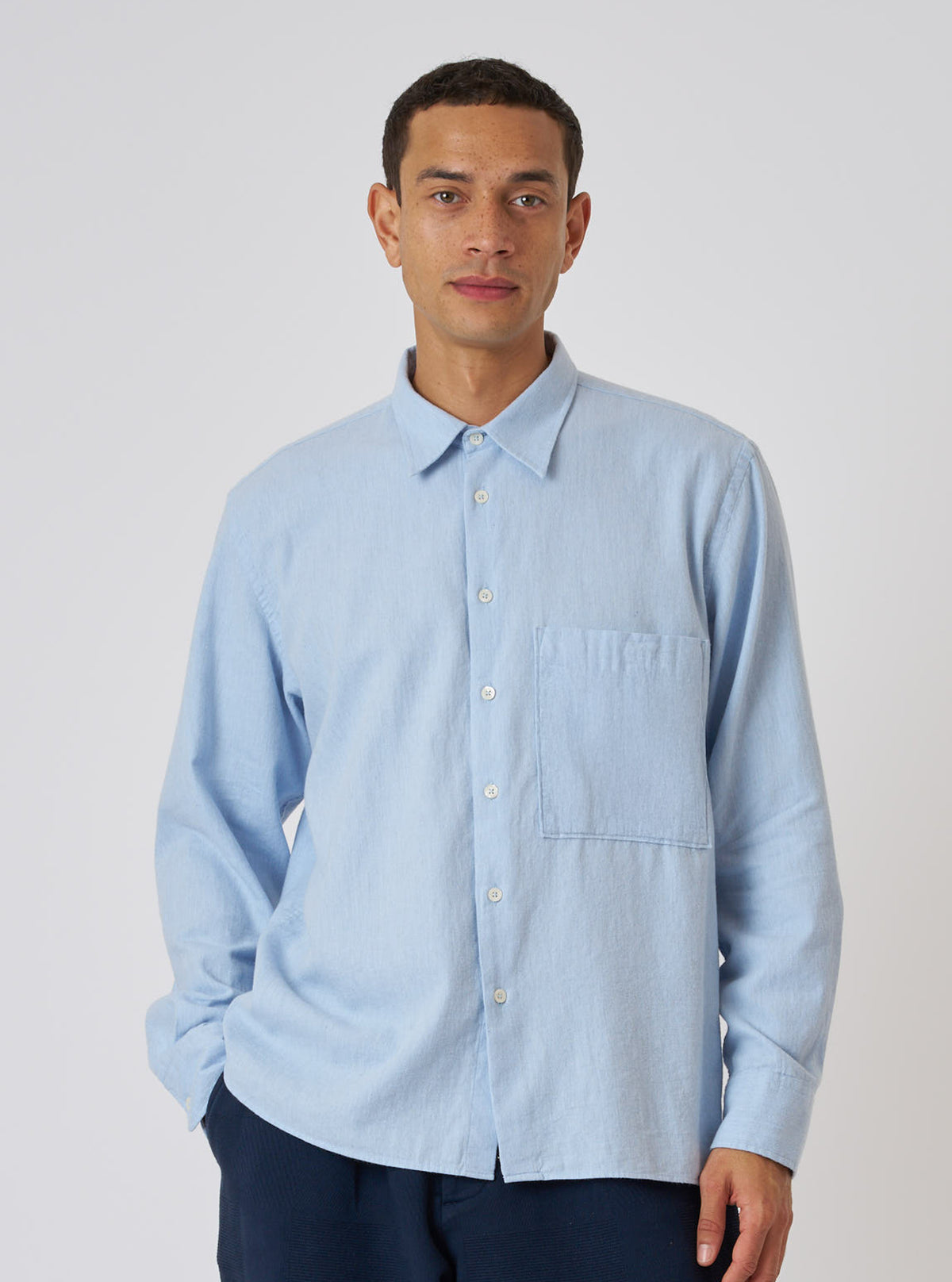 A model poses in front of a white background, wearing a Light Blue Brushed Marl Twill Square Pocket Shirt. The shirt is a light blue color and has a square pocket on the chest.