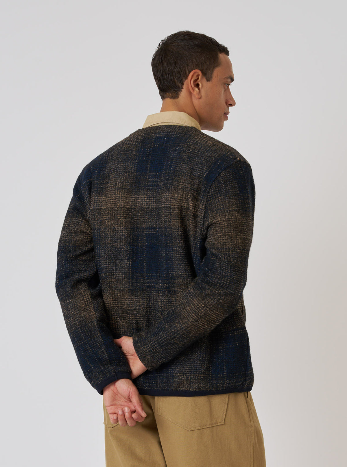 A man in a Navy Deca Check Cardigan poses against a white background. The cardigan features a navy and brown check pattern. He wears tan pants and a collared shirt.