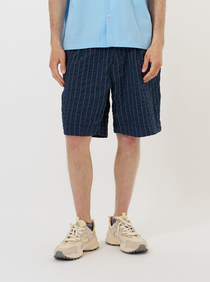 A model is wearing the Navy Chester Stripe Pleated Track Short against a plain white background. The shorts feature a navy blue and white pinstriped seersucker fabric, a relaxed long fit, and a single pleat on the front. The model is shown from the waist down, standing straight and pairing the shorts with white socks and navy blue shoes. Key details like the elasticated waistband and side pockets are clearly visible.