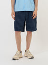 A model is wearing the Navy Chester Stripe Pleated Track Short against a plain white background. The shorts feature a navy blue and white pinstriped seersucker fabric, a relaxed long fit, and a single pleat on the front. The model is shown from the waist down, standing straight and pairing the shorts with white socks and navy blue shoes. Key details like the elasticated waistband and side pockets are clearly visible.