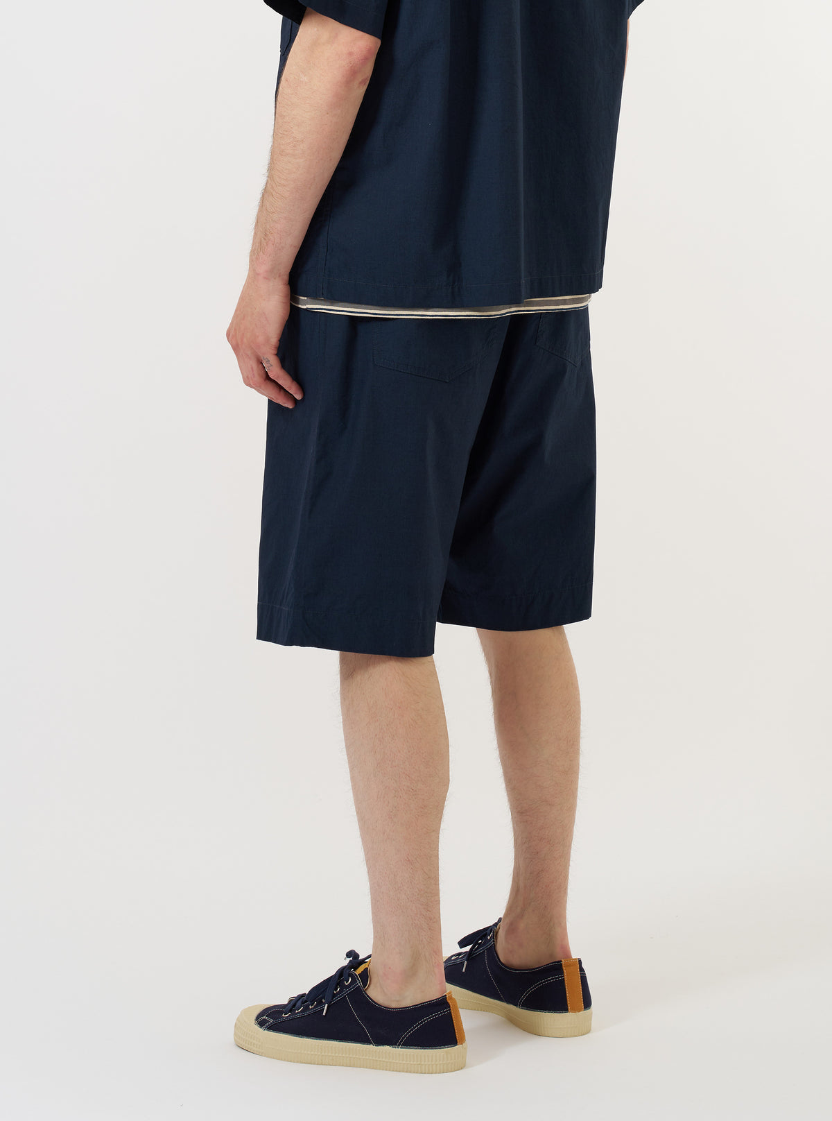 A model is wearing the Navy Paper Touch Cotton Vacation Short against a plain white background. The image, taken from a side-rear perspective, showcases the dark navy shorts with a relaxed fit and a single rear patch pocket. The model is standing and pairing the shorts with navy blue shoes. No mannequin is present in the image.