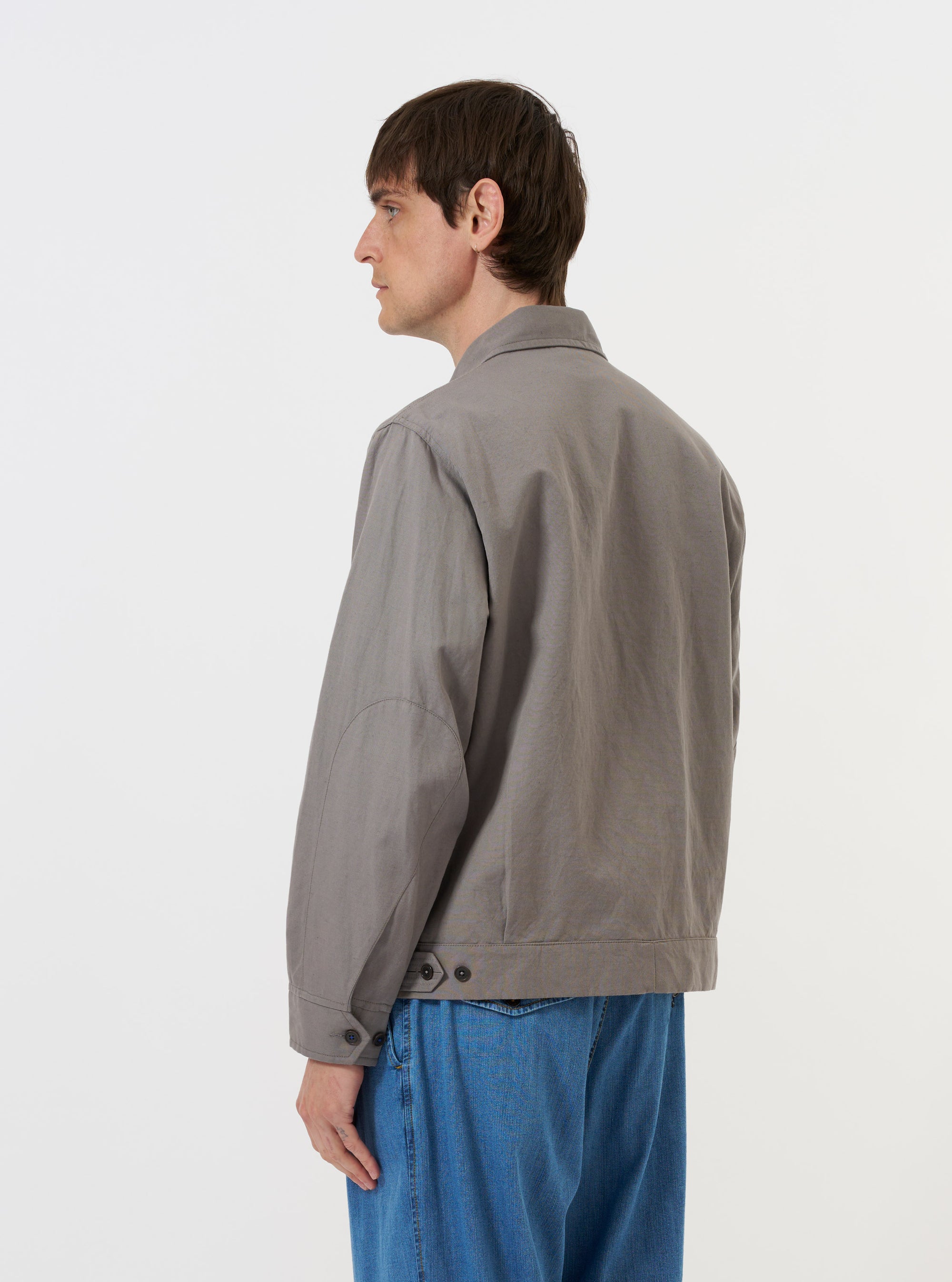 The Mid Grey Linen Slub Weave E130 Jacket is displayed flat against a plain white background. The image shows the back of the jacket, highlighting the two adjustable button tabs at the waist, reinforced elbow patches on the sleeves, and a straight hem.
