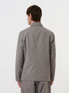 A model is wearing the Mid Grey Linen Slub Weave Bakers Jacket against a plain white background. The image provides a rear view of the jacket, highlighting its straight hem, structured shoulders, and the textured slub weave of the mid-grey fabric. The model is also wearing dark trousers.