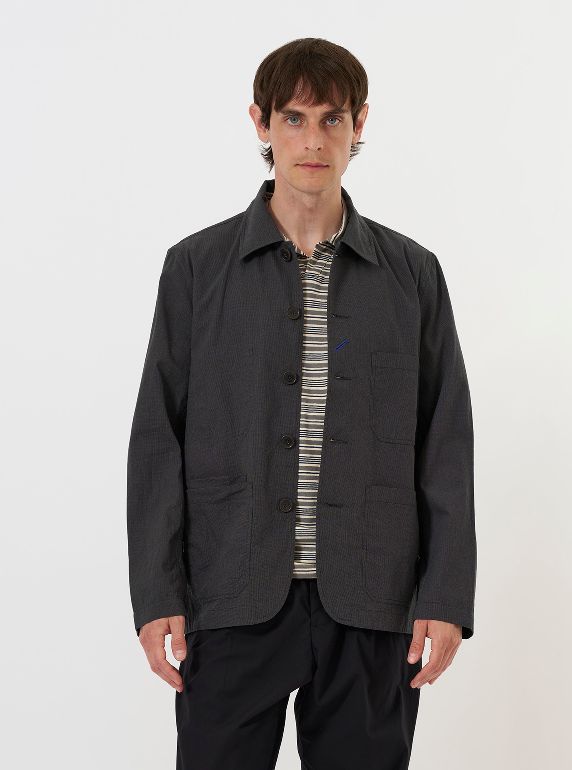 A model is wearing the Charcoal Seattle Cotton OG Bakers Jacket against a plain white background. The charcoal grey jacket is shown from the front, featuring a classic collar, a button-down front, and several large patch pockets. The model is wearing the jacket open over a light-colored top.