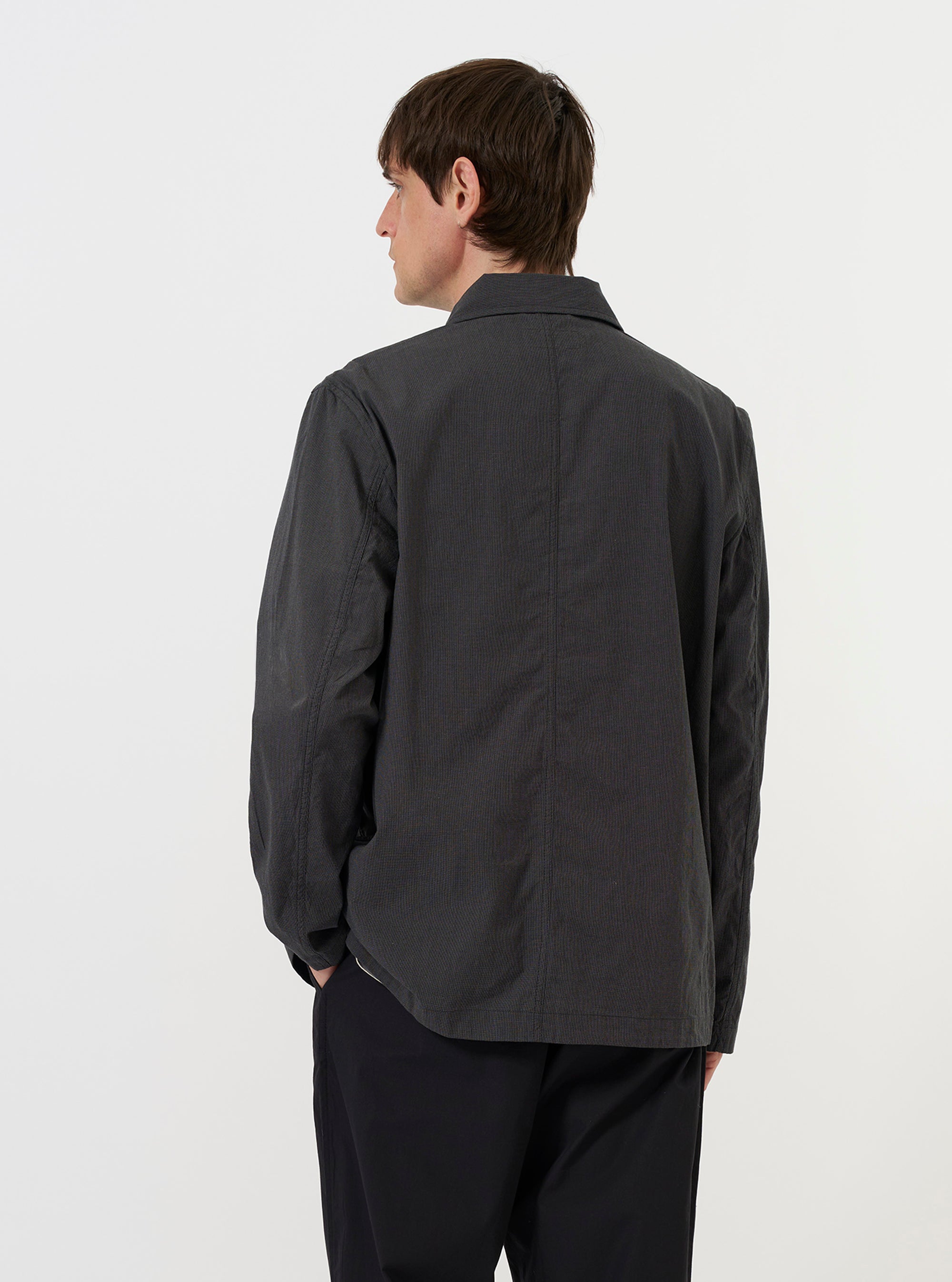 A model is wearing the Charcoal Seattle Cotton OG Bakers Jacket against a plain white background. The image, taken from a side-rear perspective, showcases the jacket's straight hem and the textured charcoal grey cotton fabric. The model's hands are placed in the large lower patch pockets. No other accessories are visible.