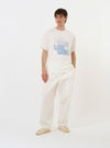 The Ecru Single Jersey Multi UW Print Tee is shown laid flat against a solid white background. The short-sleeved t-shirt features a classic crew neck and a prominent multi-colored graphic print centered on the chest, consisting of four graduated rows of the letters "UW" in shades of blue, orange, and navy.