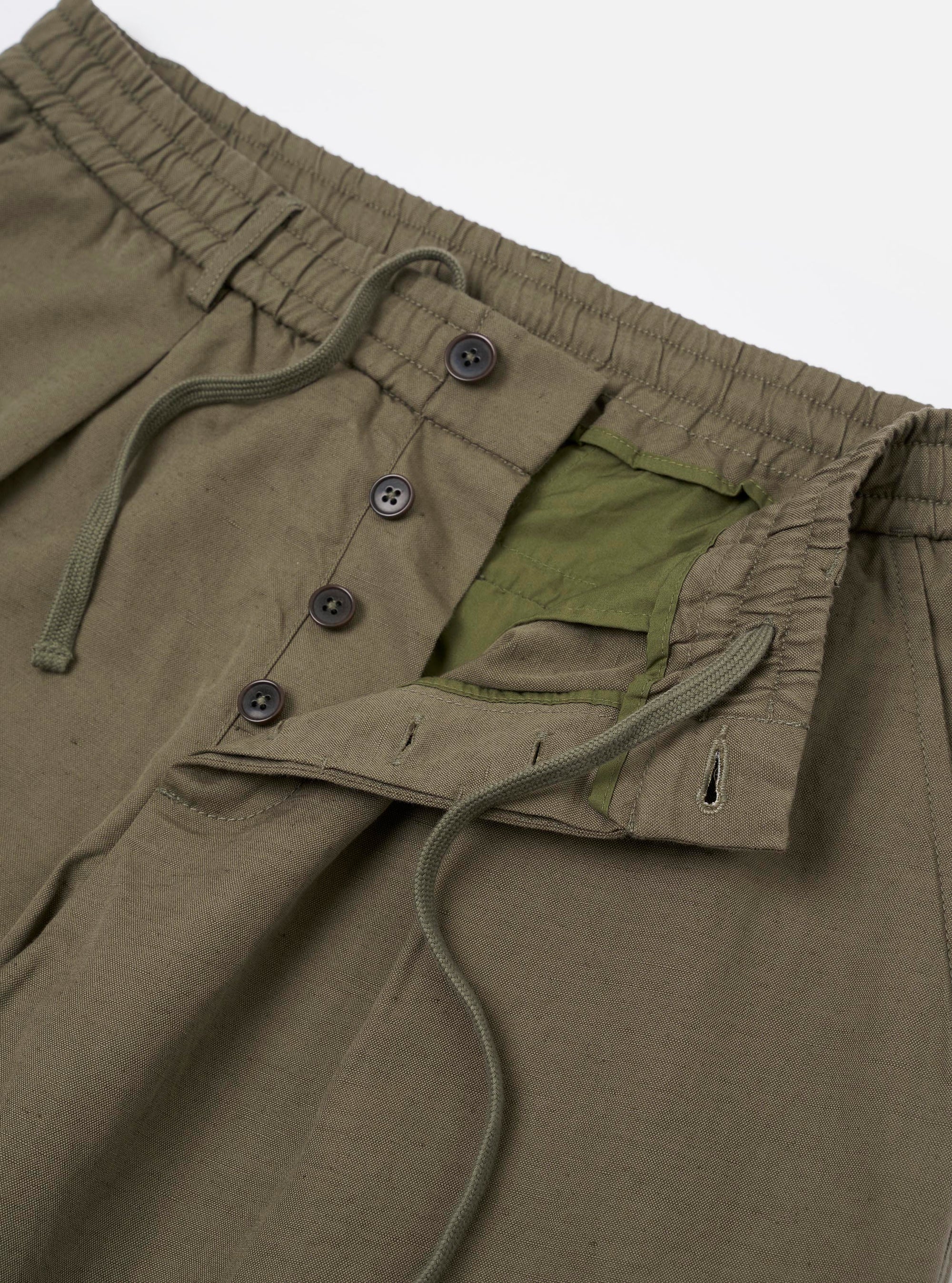A pair of olive green linen slub weave Oxford pants is shown from a rear three-quarter angle, laid flat against a plain white background. The image highlights the relaxed, wide-leg fit and the textured appearance of the slub fabric. A single rear patch pocket with a button closure is visible on the right side.