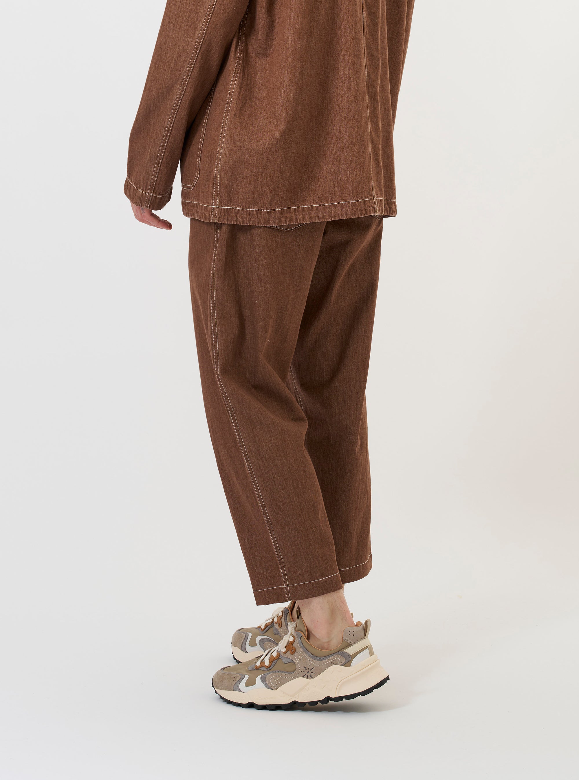A model wears the Brown Meco Denim Hi Water Trouser against a plain white background. The trousers are a slim, cropped fit made from brown denim fabric, featuring a visible drawstring at the waistband and side pockets. The image focuses on the lower half of the model's body to highlight the fit and texture of the garment.