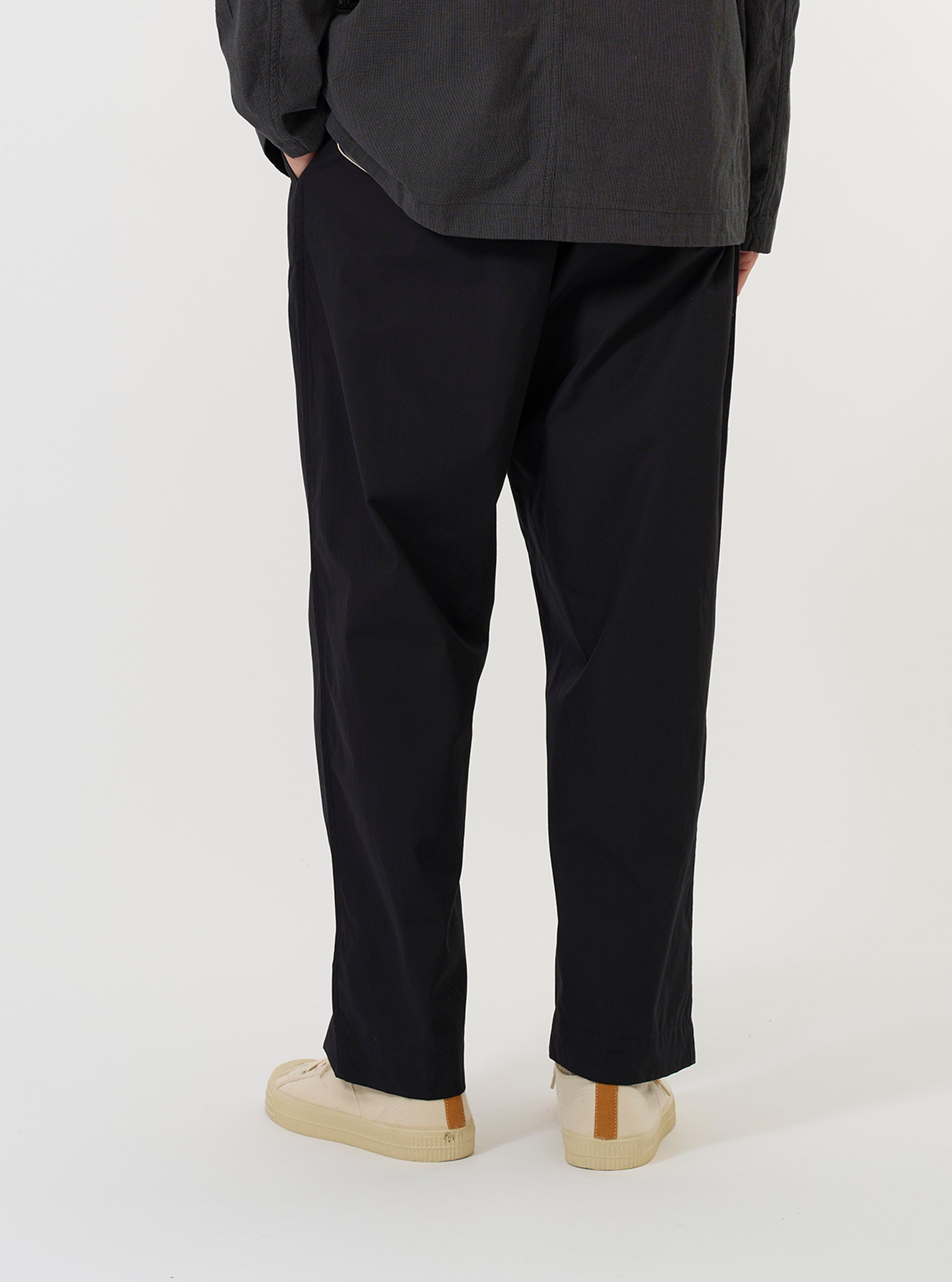 A model is wearing the Black Broadcloth Oxford Pant against a plain white background. The image provides a view from the waist down, showing the loose, wide-leg fit of the black trousers, featuring a pleated front and a drawstring waistband.