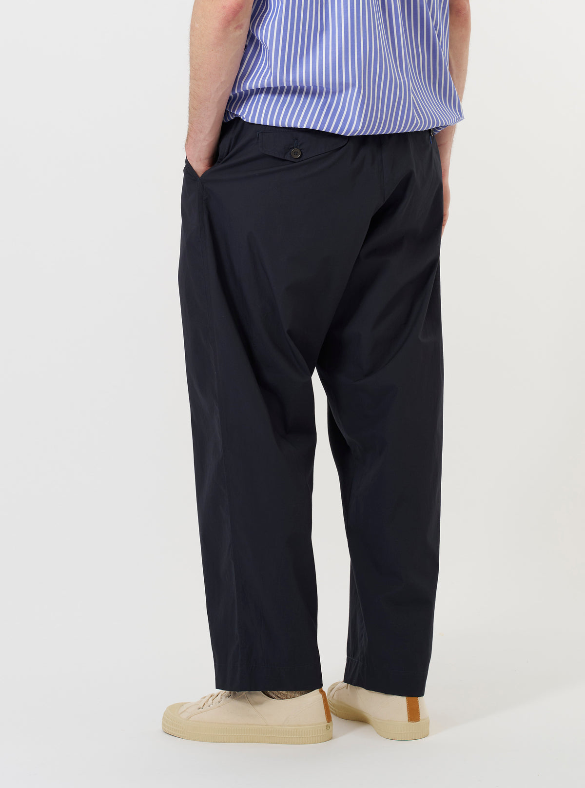 A model is wearing the Navy Broadcloth Oxford Pant against a plain white background. The image provides a view of the trousers from the waist down, showcasing the loose, wide fit and the deep navy blue color of the broadcloth fabric. The model is standing, and the trousers drape naturally, highlighting the pleated front and the textured finish of the material.