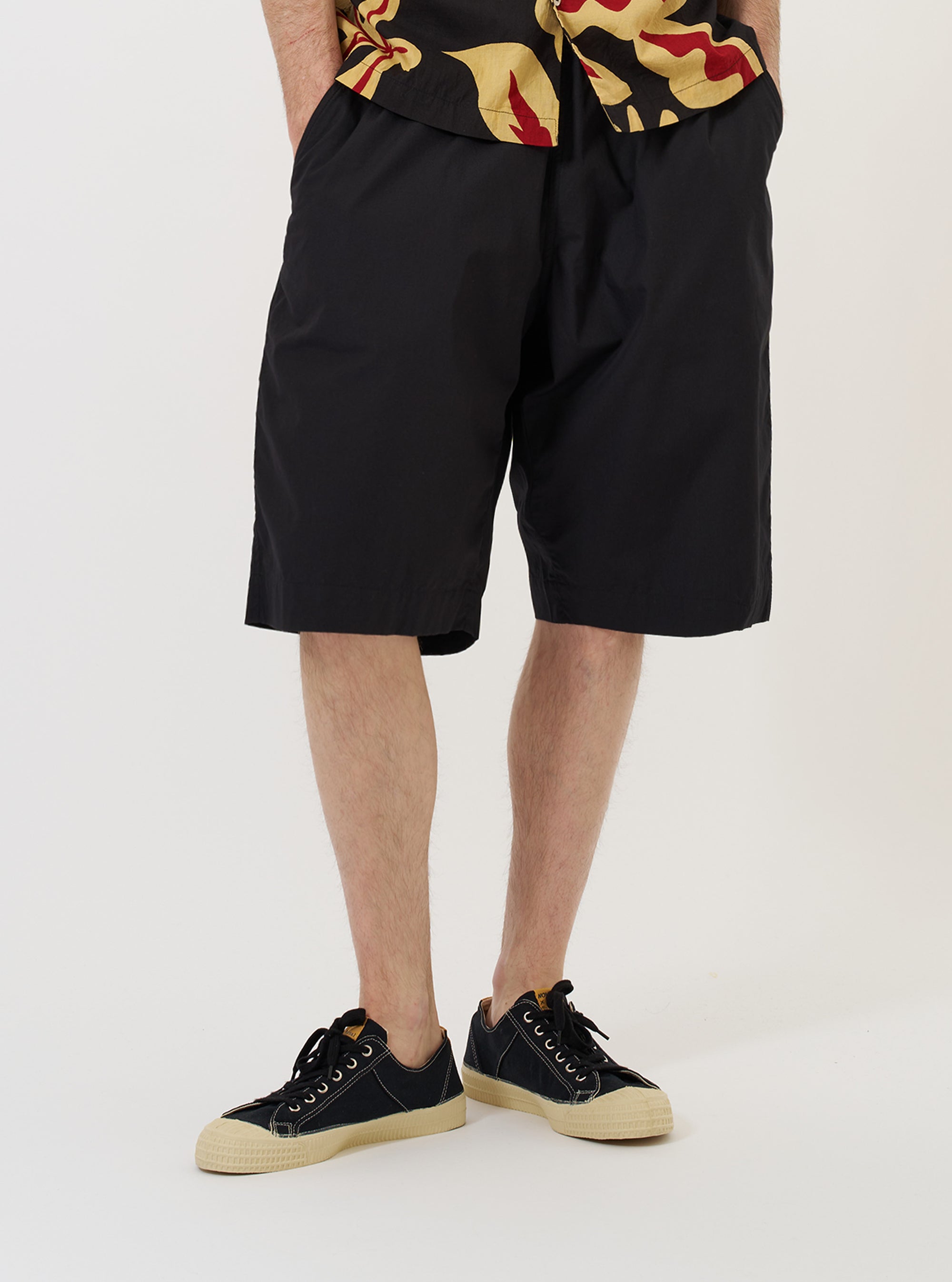 A model stands against a plain white background wearing the Black Broadcloth Vacation Short. The image captures the garment from the waist down, highlighting the relaxed fit, mid-thigh length, and the smooth texture of the black cotton broadcloth. The model is standing straight and pairing the shorts with dark-colored shoes and white socks. No mannequin is present in the image.