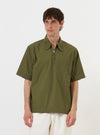 A model is wearing the Olive Broadcloth Ewan Shirt against a plain white background. The olive green shirt is shown from the torso up, featuring a relaxed, boxy fit, short sleeves, and a visible 1/4 zip closure at the neckline. The garment is worn over the model's upper body, highlighting the smooth texture of the broadcloth fabric.