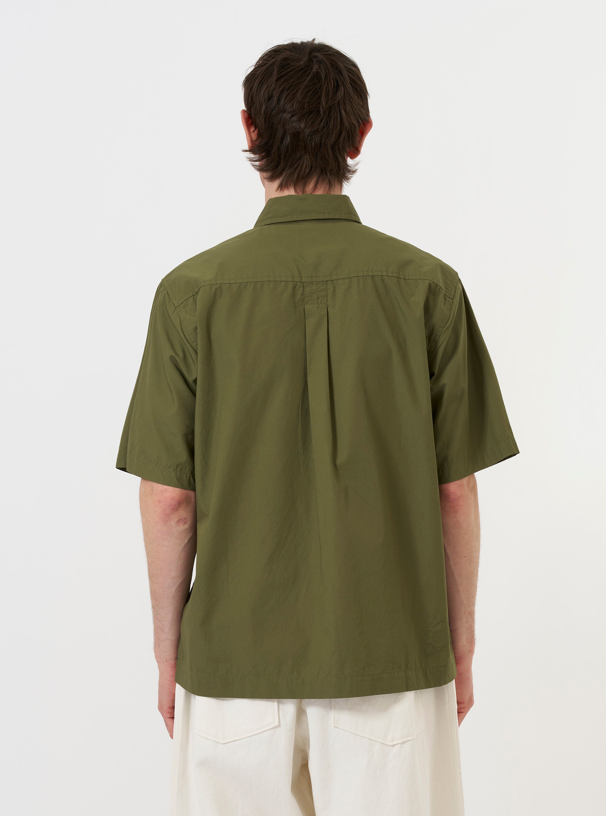 A model is wearing the Olive Broadcloth Ewan Shirt against a plain white background. The image provides a rear view of the model from the waist up, highlighting the olive green fabric, the back yoke, and the straight hem of the garment. The model is also wearing dark trousers. No mannequin is present in the image.