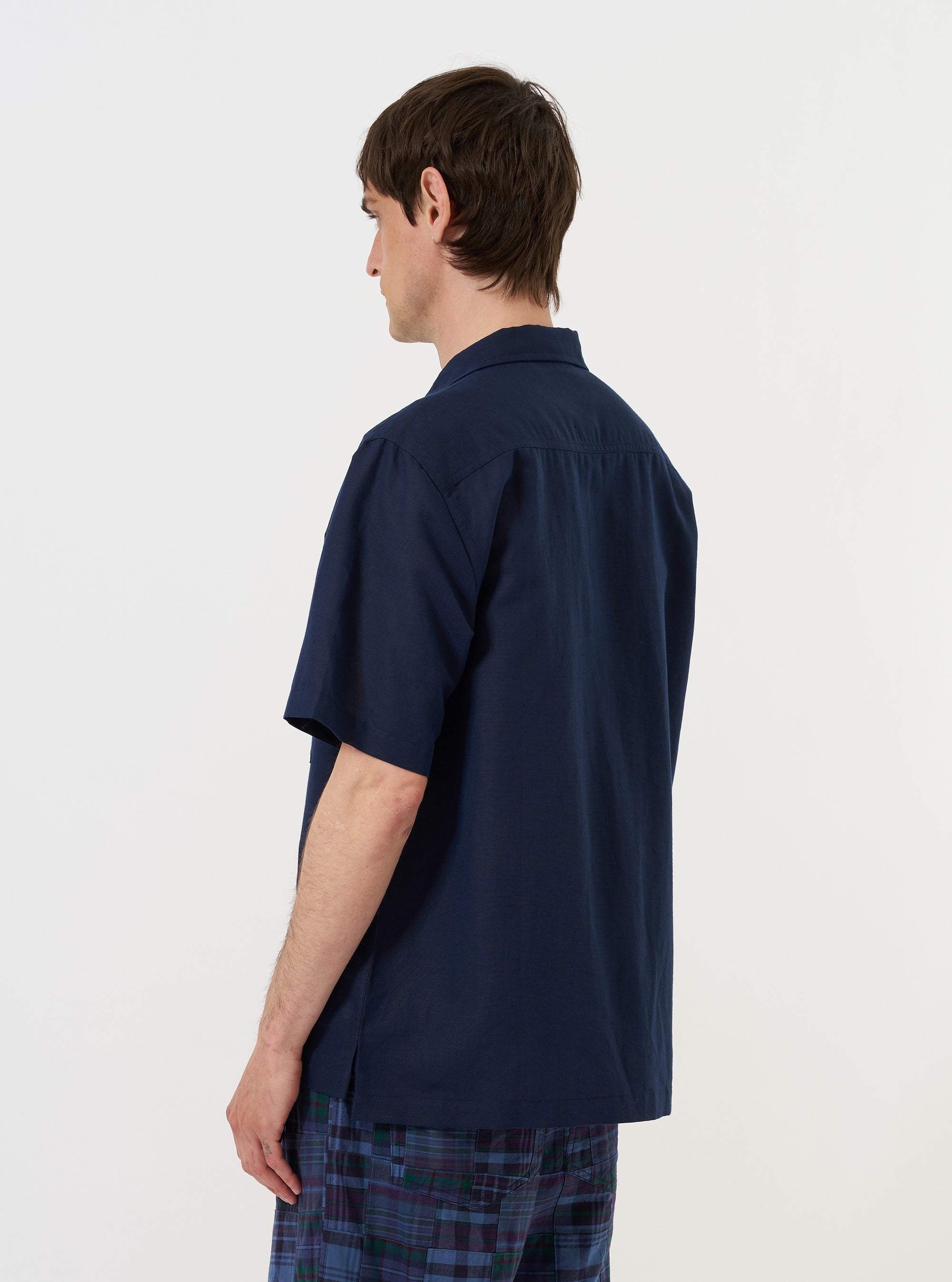 A model is wearing the Navy Linen Cotton Shirting Camp Shirt against a plain white background. The image provides a rear view of the model from the waist up, highlighting the shirt's back yoke with subtle pleats and the relaxed, loose fit of the short sleeves. The navy blue fabric has a slightly textured appearance, and the model is shown with their arms at their sides.