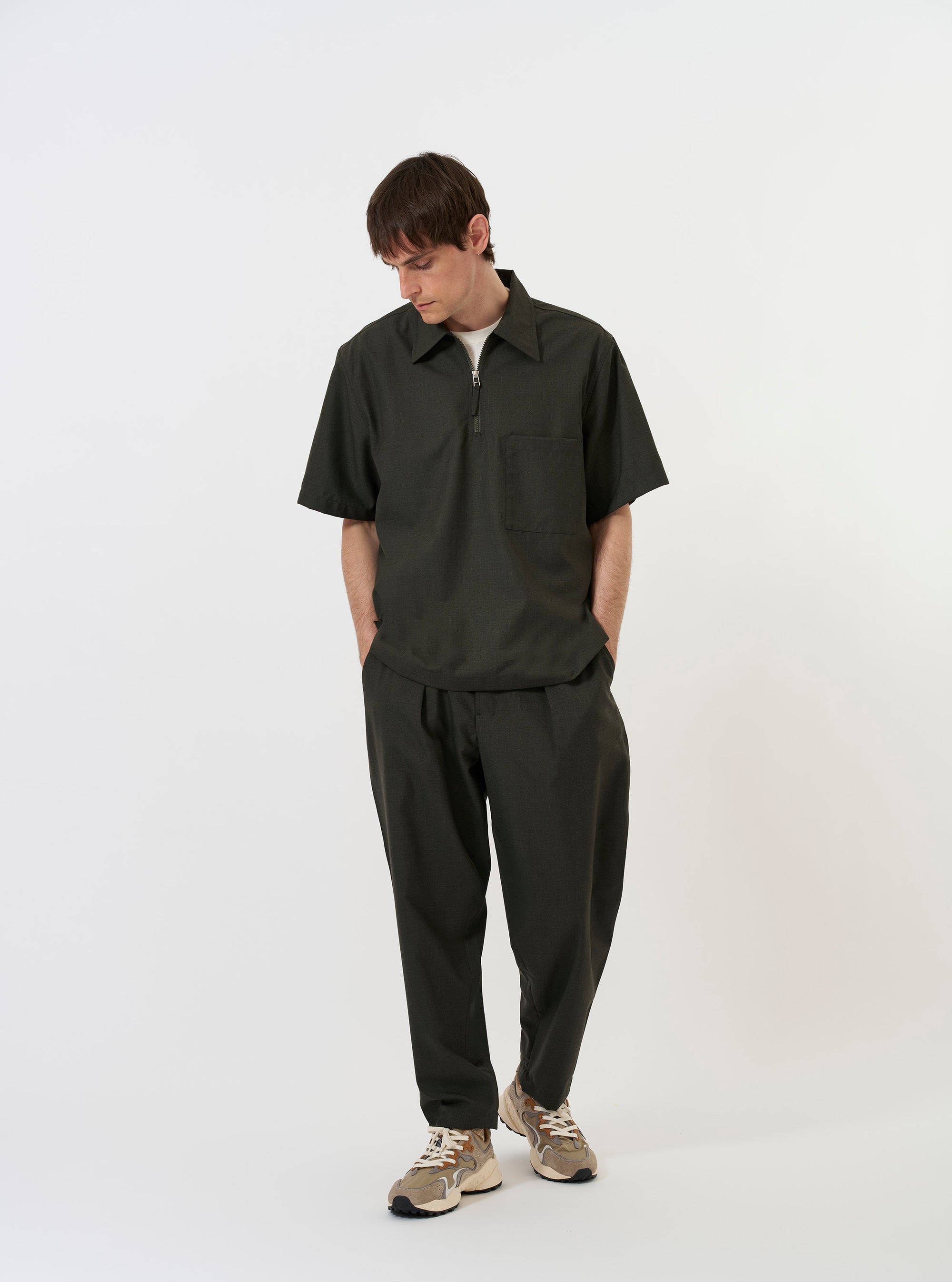The Olive Marl Tropical Suiting Ewan Shirt is shown laid flat against a solid white background. The garment is a short-sleeved, pullover-style shirt featuring a half-zip front, a classic pointed collar, and a straight hem. The fabric has a subtle olive green marl texture.