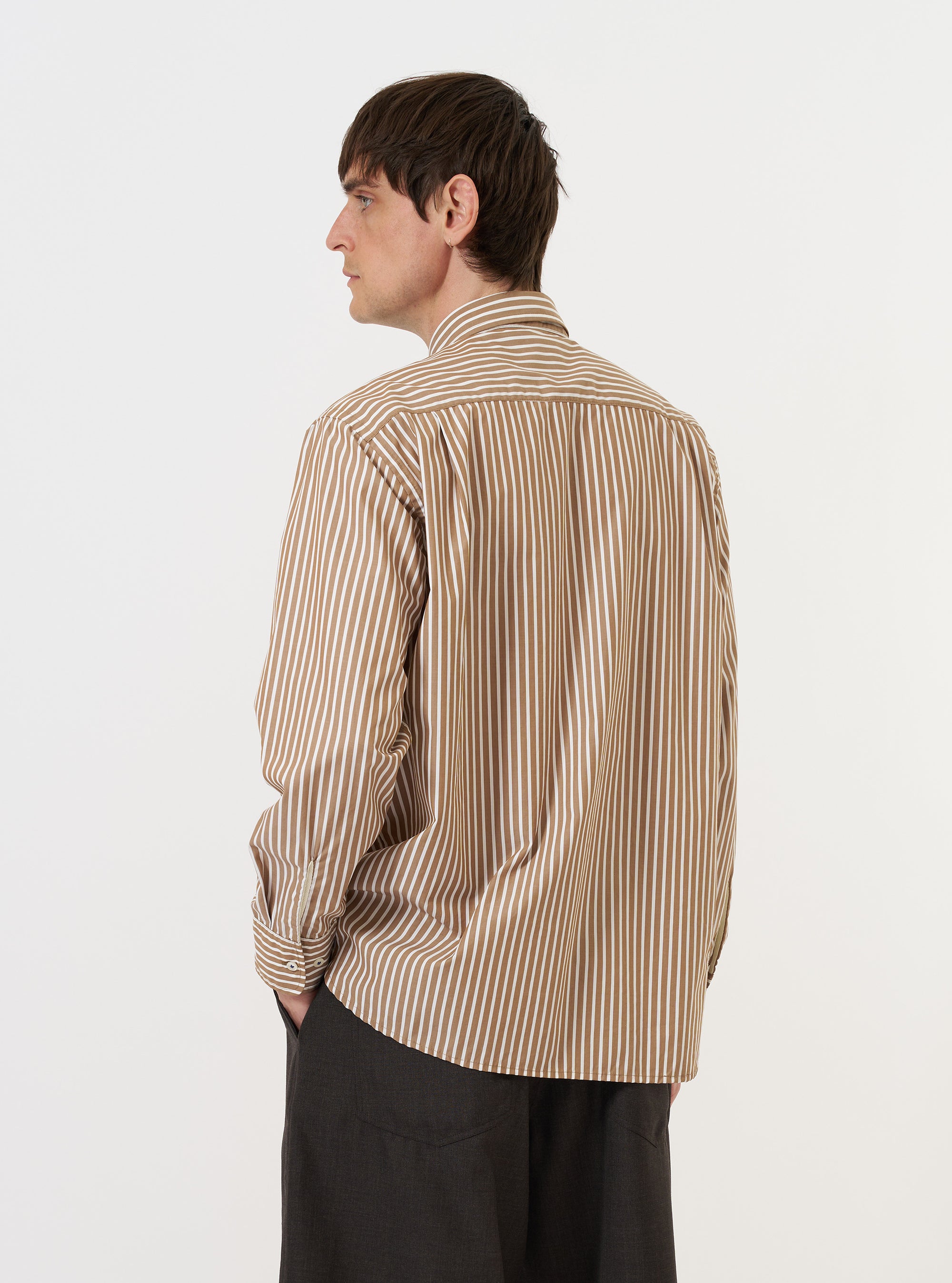 A model is wearing the Brown/White Cotton Stripe Square Pocket Shirt against a plain white background. The image shows the shirt from the back, highlighting the clean back yoke, long sleeves, and the fine brown and white vertical stripe pattern of the cotton fabric. The model is wearing the shirt untucked, paired with dark trousers.