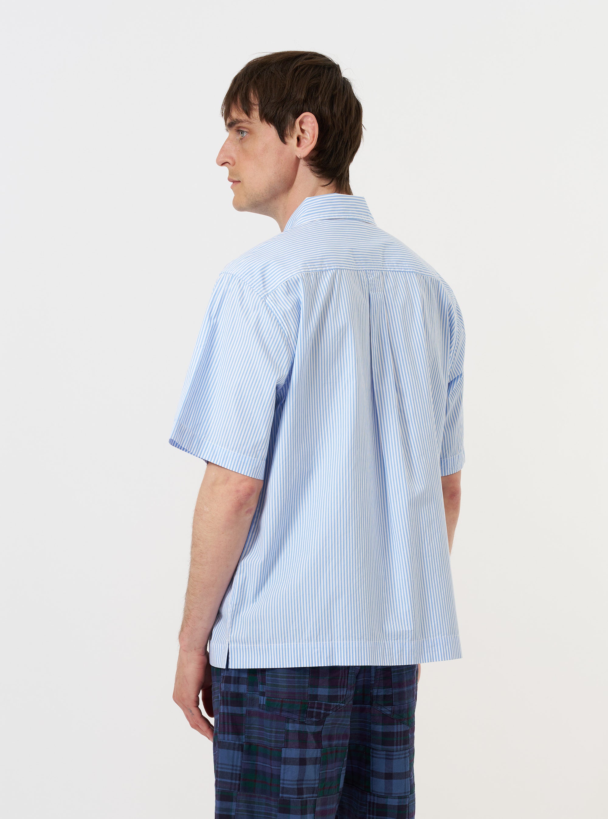 A model is wearing the Sky/White Classic Nipon Stripe Ewan Shirt against a plain white background. Seen from the back, the short-sleeved shirt features vertical sky blue and white stripes, a horizontal back yoke, and a central box pleat. The model is wearing the shirt untucked.
