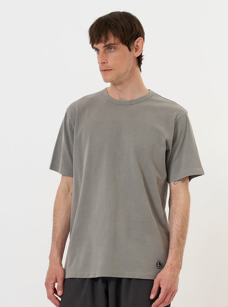 A model is wearing the Mid Grey Piquet Core Tee while standing against a plain white background. The mid-grey short-sleeved t-shirt features a subtle textured piquet weave and a ribbed crew neckline. The model is shown from the chest up, highlighting the fit and fabric texture of the garment.