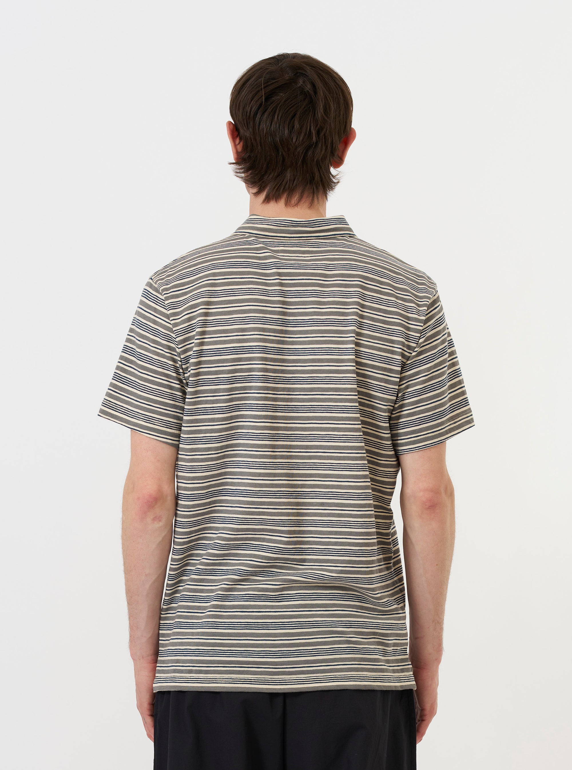 A model stands against a plain white background wearing the Grey OS Stripe Jersey Vacation Polo. The image shows the garment from the back, highlighting the grey and white horizontal stripes, the straight hem, and the relaxed fit of the short-sleeved polo shirt.
