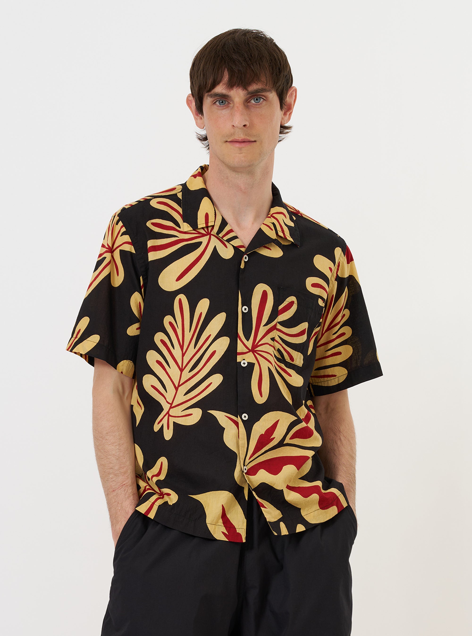 A model stands against a plain white background wearing the Black Baja Print Road Shirt. The short-sleeved shirt features a lightweight black cotton fabric with a white Baja-inspired geometric print. It is styled with the top button open to reveal a single-piece camp collar and has a visible patch pocket on the left chest. The model is shown from the waist up.