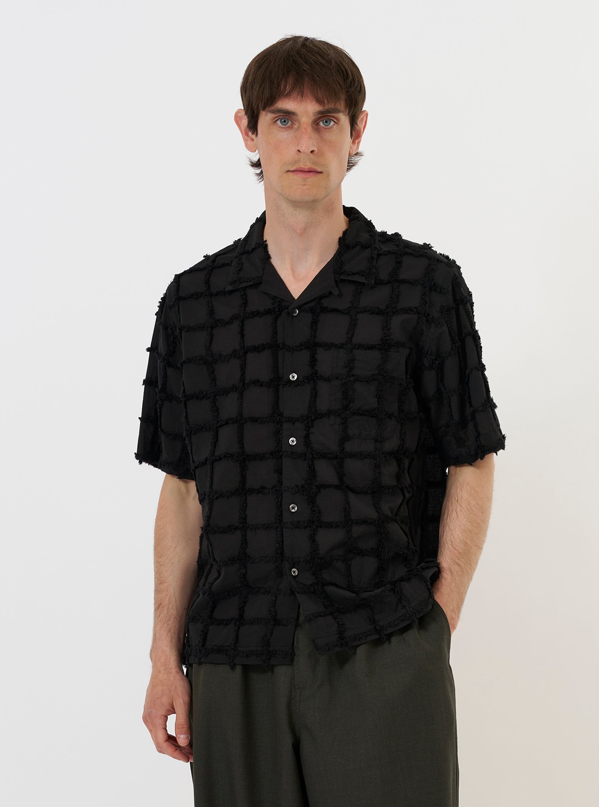 A model is wearing the Black 3D Check Road Shirt against a plain white background. The shirt features a textured black and grey 3D check pattern, a relaxed camp collar, and a button-down front. The model is shown from the waist up, wearing the shirt open over a dark undershirt, highlighting the garment's fit and the intricate weave of the fabric.