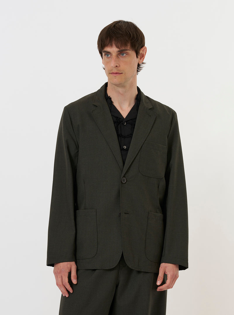 A model stands against a plain white background wearing the Olive Marl Tropical Suiting Two Button Jacket. The jacket is an unstructured, relaxed fit in olive green marl, featuring a two-button front closure, a long, wide lapel, two lower patch pockets, and a single chest patch pocket. The model is shown from the waist up, layering the jacket over a simple top.