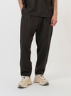 A model is wearing the Brown Marl Tropical Suiting Pleated Track Pant against a plain white background. The trousers are shown from the waist down, featuring a loose and tapered fit with a pleated front and a visible drawstring at the waistband. The brown marl fabric has a subtle textured appearance. The model is standing straight, pairing the trousers with dark-colored shoes.