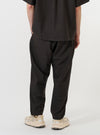 A model is wearing the Brown Marl Tropical Suiting Pleated Track Pant against a plain white background. The image, captured from a side-rear perspective, showcases the loose, tapered fit and the brown marl texture of the lightweight tropical suiting fabric. The model is standing, highlighting the drape of the trousers and the narrow hem at the ankle.