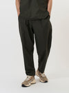 A model is wearing the Olive Marl Tropical Suiting Pleated Track Pant against a plain white background. The image shows the model from the waist down, highlighting the loose, tapered fit of the olive green trousers. Key details visible include the single pleat at the front, side slip pockets, and the drape of the tropical suiting fabric. The model is standing and is also wearing dark shoes.