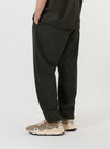 A model is shown from the waist down wearing the Olive Marl Tropical Suiting Pleated Track Pant against a plain white background. The image provides a side-rear perspective of the olive green trousers, highlighting the loose, tapered fit and the texture of the tropical suiting fabric. The model is standing, showcasing the relaxed drape of the garment.