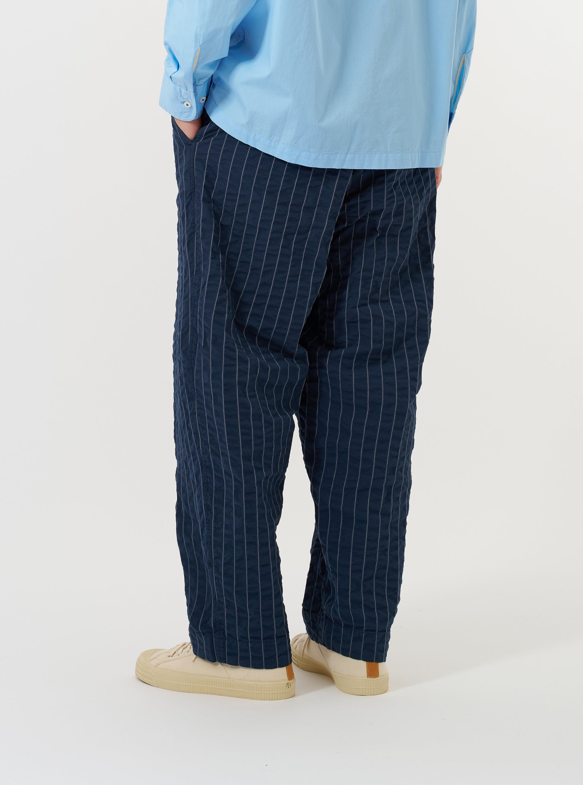 A model is wearing the Navy Chester Stripe Oxford Pant against a plain white background. The image, taken from a side-rear perspective, showcases the loose, wide-leg fit of the navy blue trousers, which feature thin vertical white stripes and a single rear buttoned pocket.