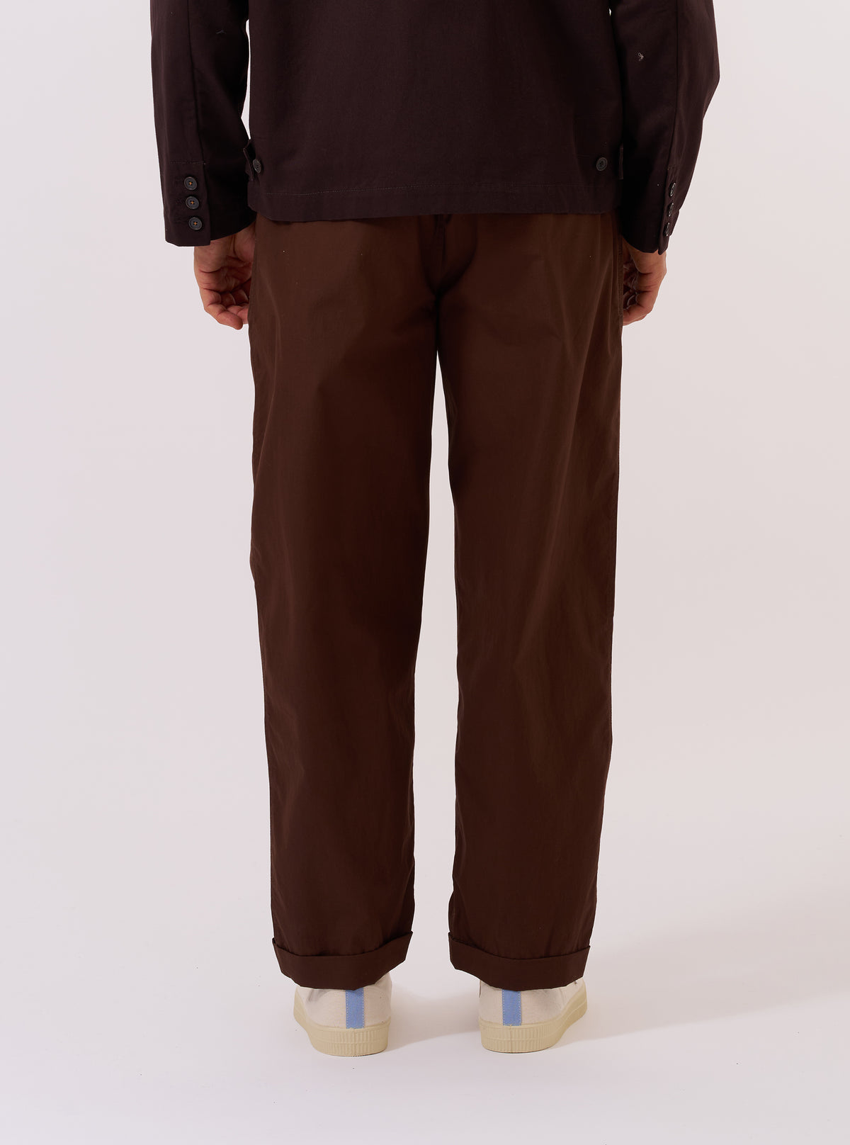 A model wears the Paraboot x Universal Works Brown Peached Poplin Super Chino, shown from a rear three-quarter angle against a plain white background. The image highlights the relaxed, straight-leg fit of the trousers and the smooth, matte texture of the brown peached poplin fabric. The model is wearing the chinos with dark shoes and a white T-shirt.