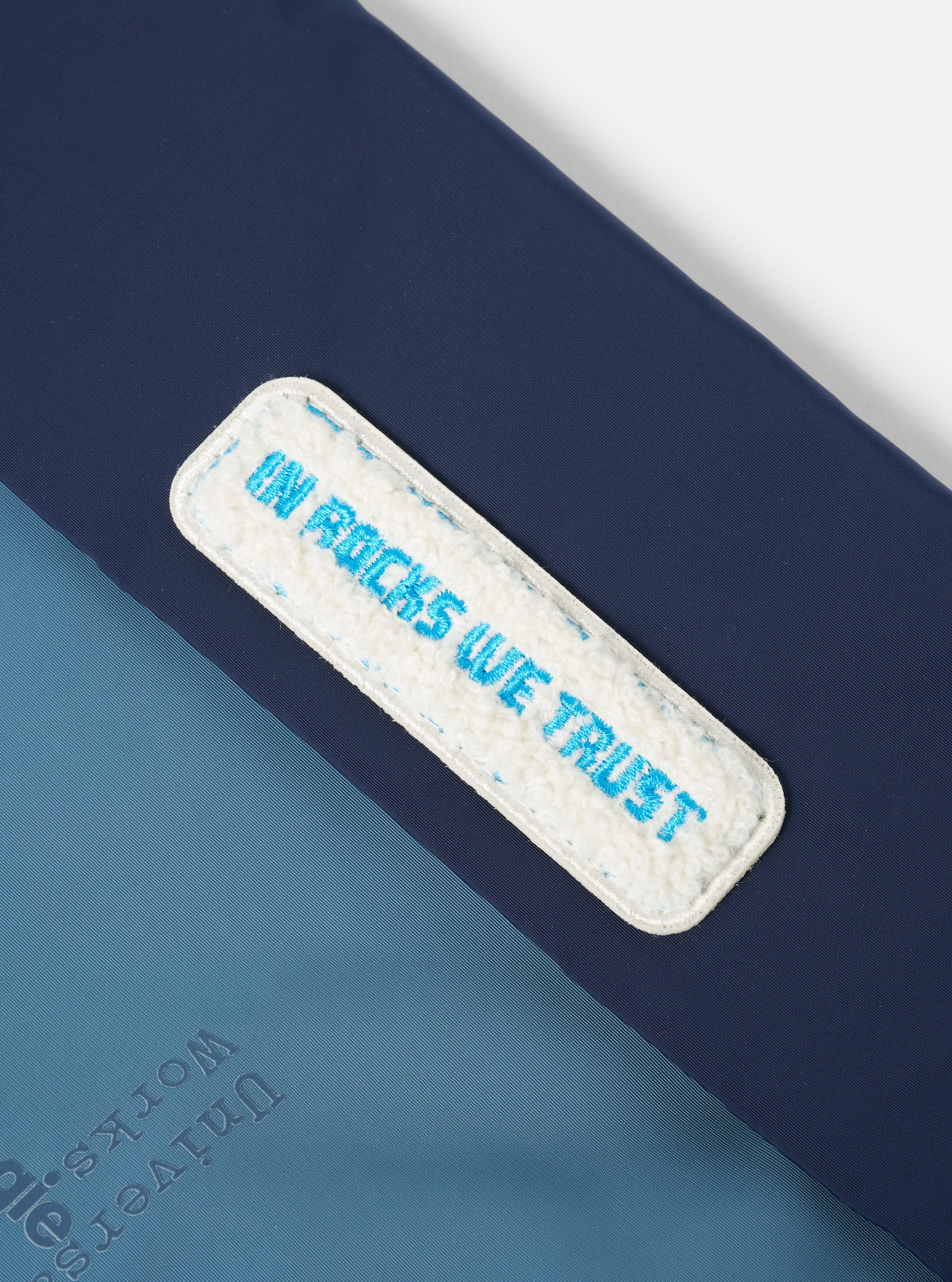 Close-up of the Topologie x Universal Works Insignia Blue UW Tech Musette Medium. Features a blue tech material with a white patch that reads "IN ROCKS WE TRUST" in blue lettering.