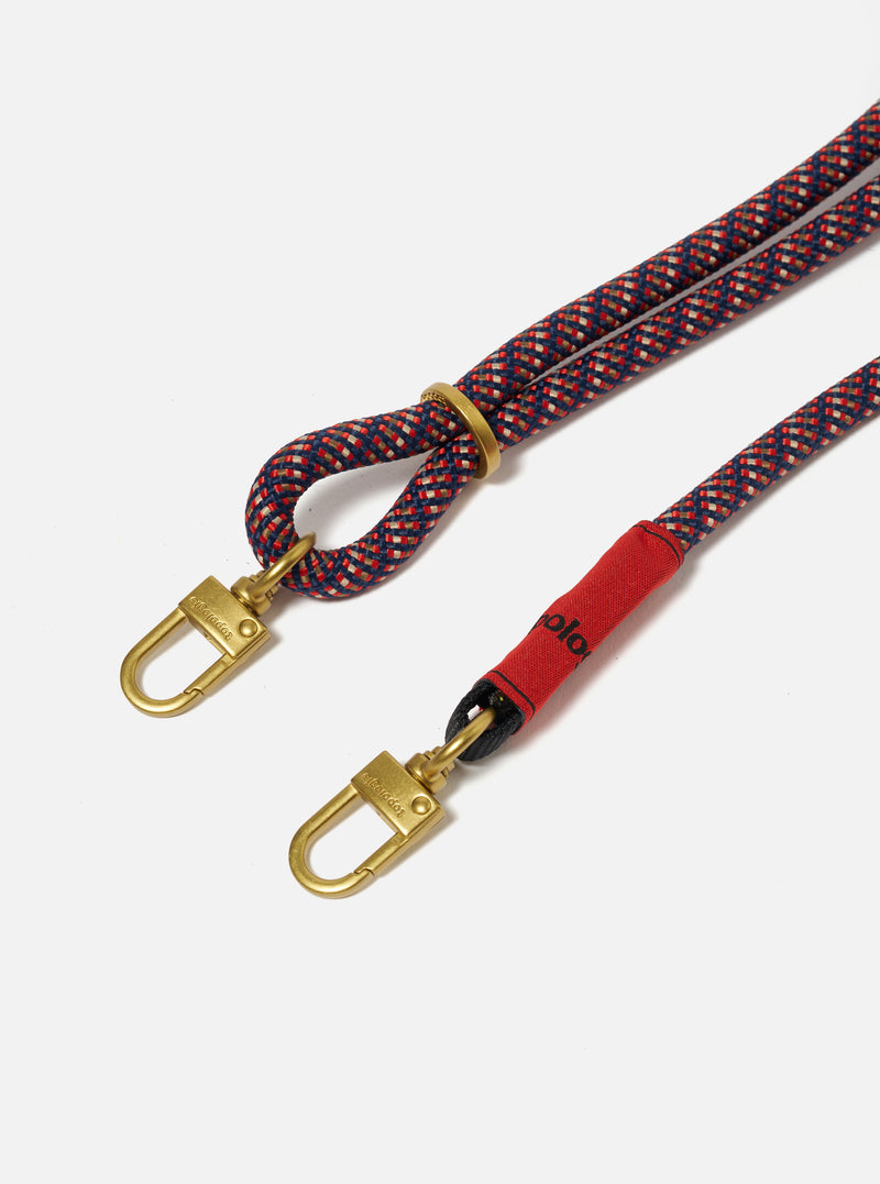 Close-up of the Topologie x Universal Works Olive Hitch UW Rope 8mm Strap on a white background. The rope is red, blue and beige with gold hardware and a red fabric section.