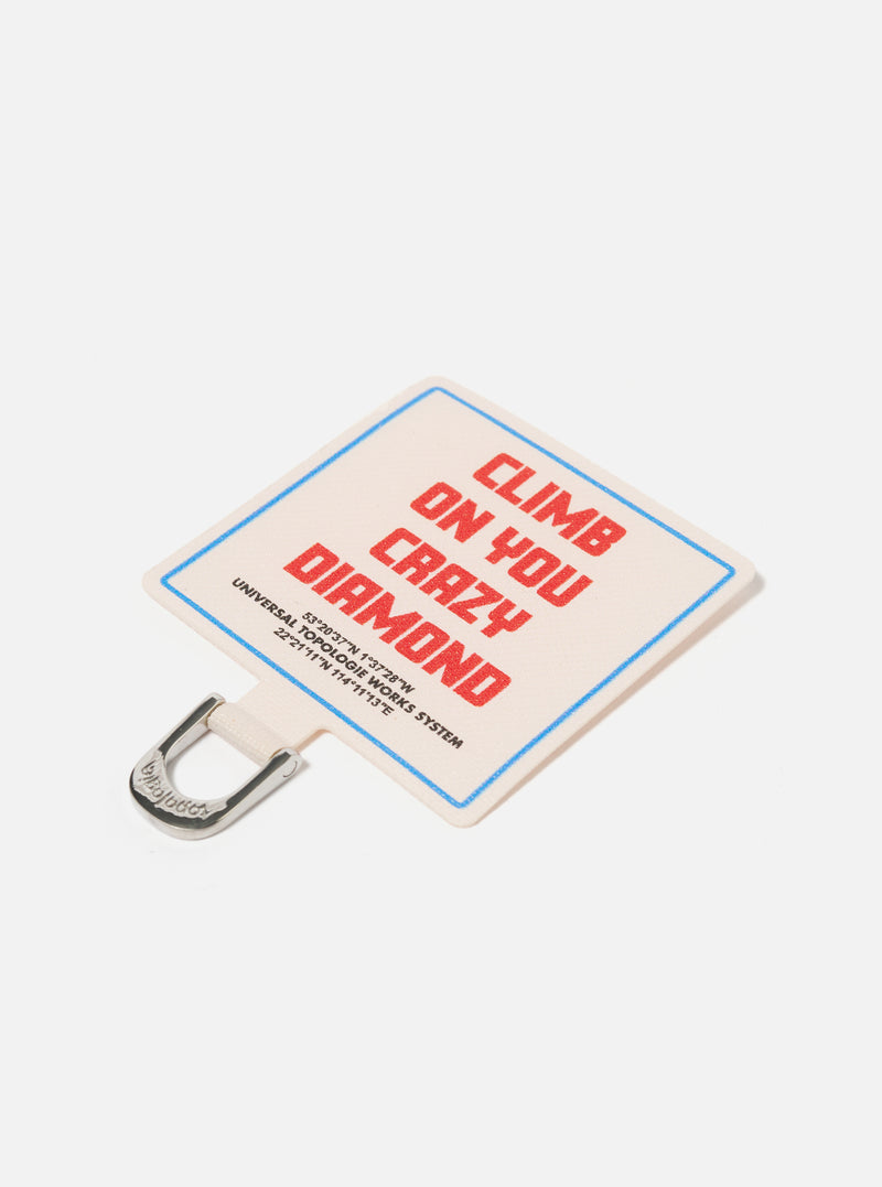 Close-up of the Topologie x Universal Works Off White UW Tech Phone Strap Adapter. It's off-white with red text and a blue border, featuring a silver metal clip. Shot on a clean white background.