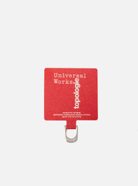 Close-up of the Topologie x Universal Works Red UW Tech Phone Strap Adapter on a white background. The red adapter features the logos of both brands.