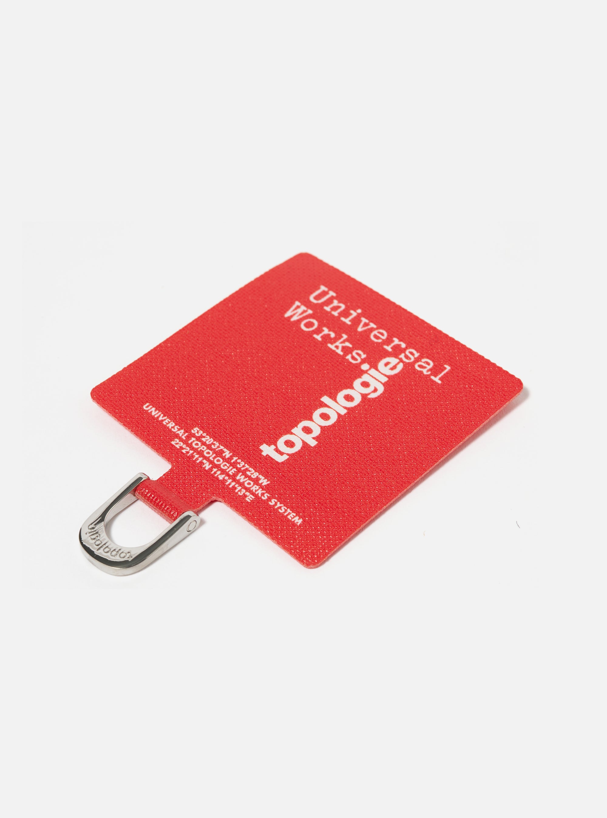 Close-up of the Topologie x Universal Works Red UW Tech Phone Strap Adapter on a white surface. The red fabric adapter features white text and a silver metal loop.