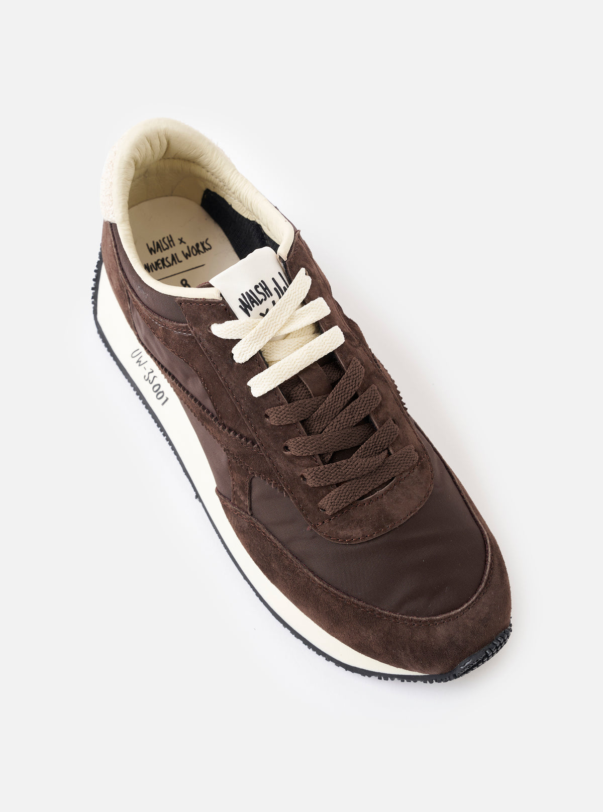 An angled three-quarter view of the pair of Walsh x Universal Works Tornado sneakers in brown leather and canvas on a white background, highlighting the lace-up closure, tonal stitching, textured textile panels, and durable rubber soles.