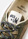A model wearing the Walsh x Universal Works Ensign in Green Leather/Canvas sneakers shown from the waist down in a clean studio environment, presenting the shoes styled naturally with modern relaxed-fit trousers.