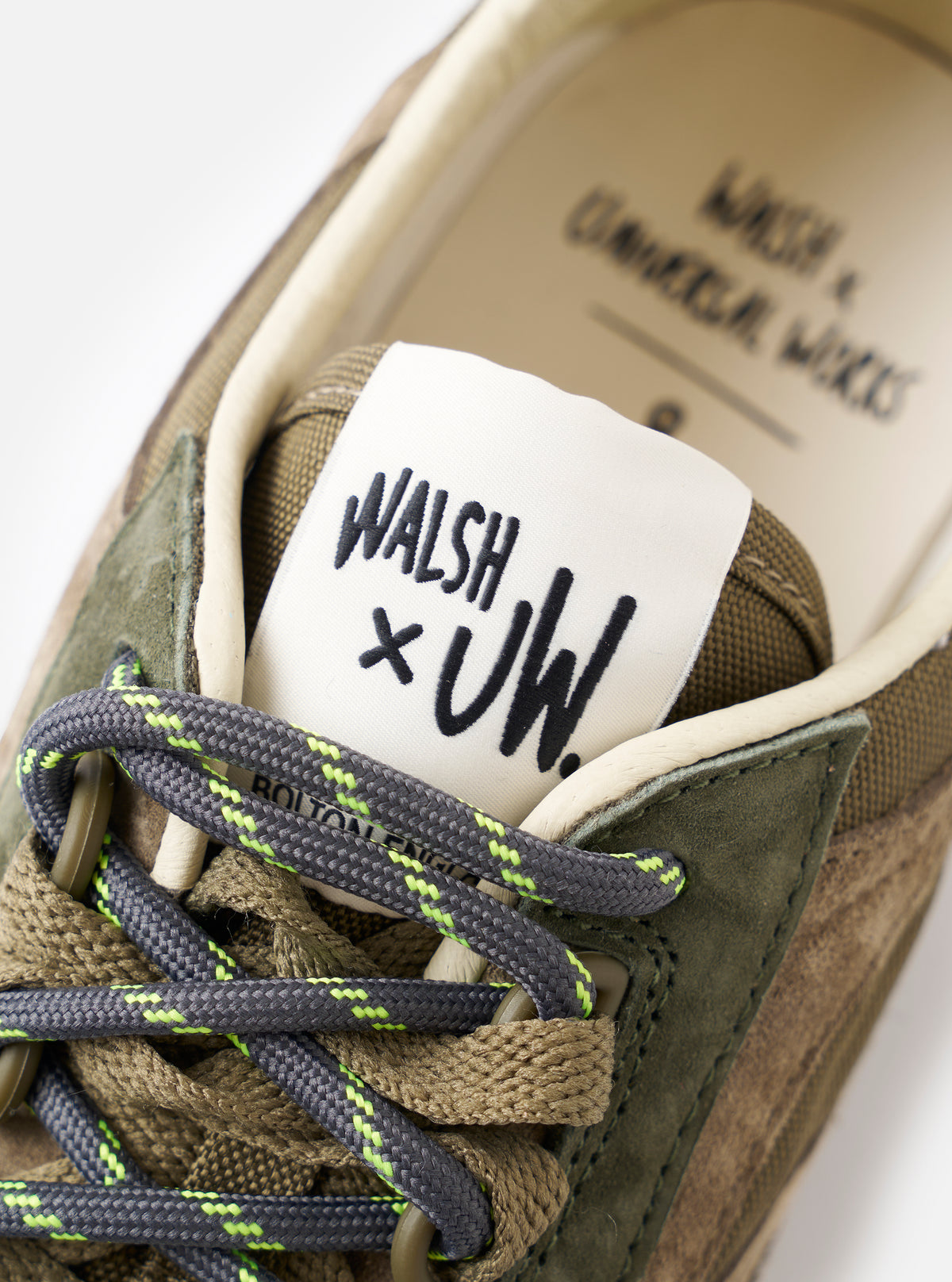 A model wearing the Walsh x Universal Works Ensign in Green Leather/Canvas sneakers shown from the waist down in a clean studio environment, presenting the shoes styled naturally with modern relaxed-fit trousers.
