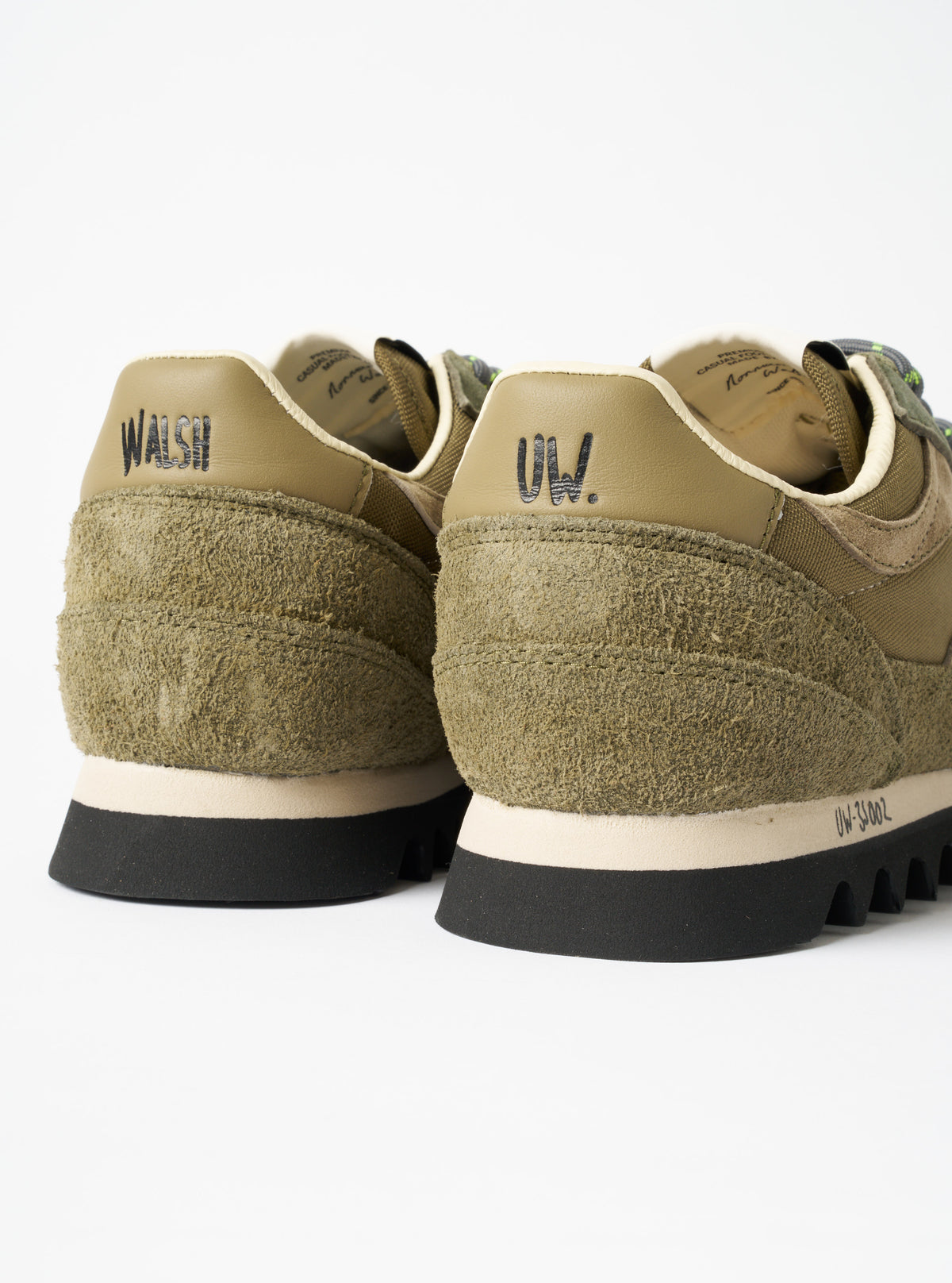 A model wearing the Walsh x Universal Works Ensign in Green Leather/Canvas sneakers styled with dark trousers, photographed against a plain neutral background to demonstrate real-world fit.