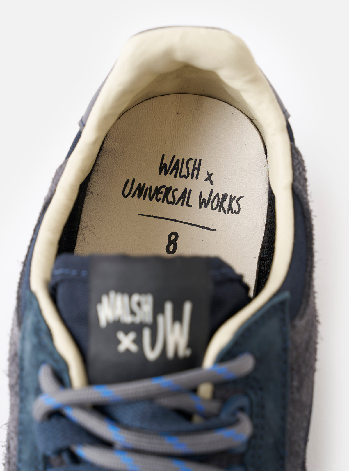 A close-up detail shot of the Walsh x Universal Works Ensign in Navy Leather/Canvas sneaker against a white studio background, highlighting the contrast stitching, suede overlays, and metallic D-ring eyelets.