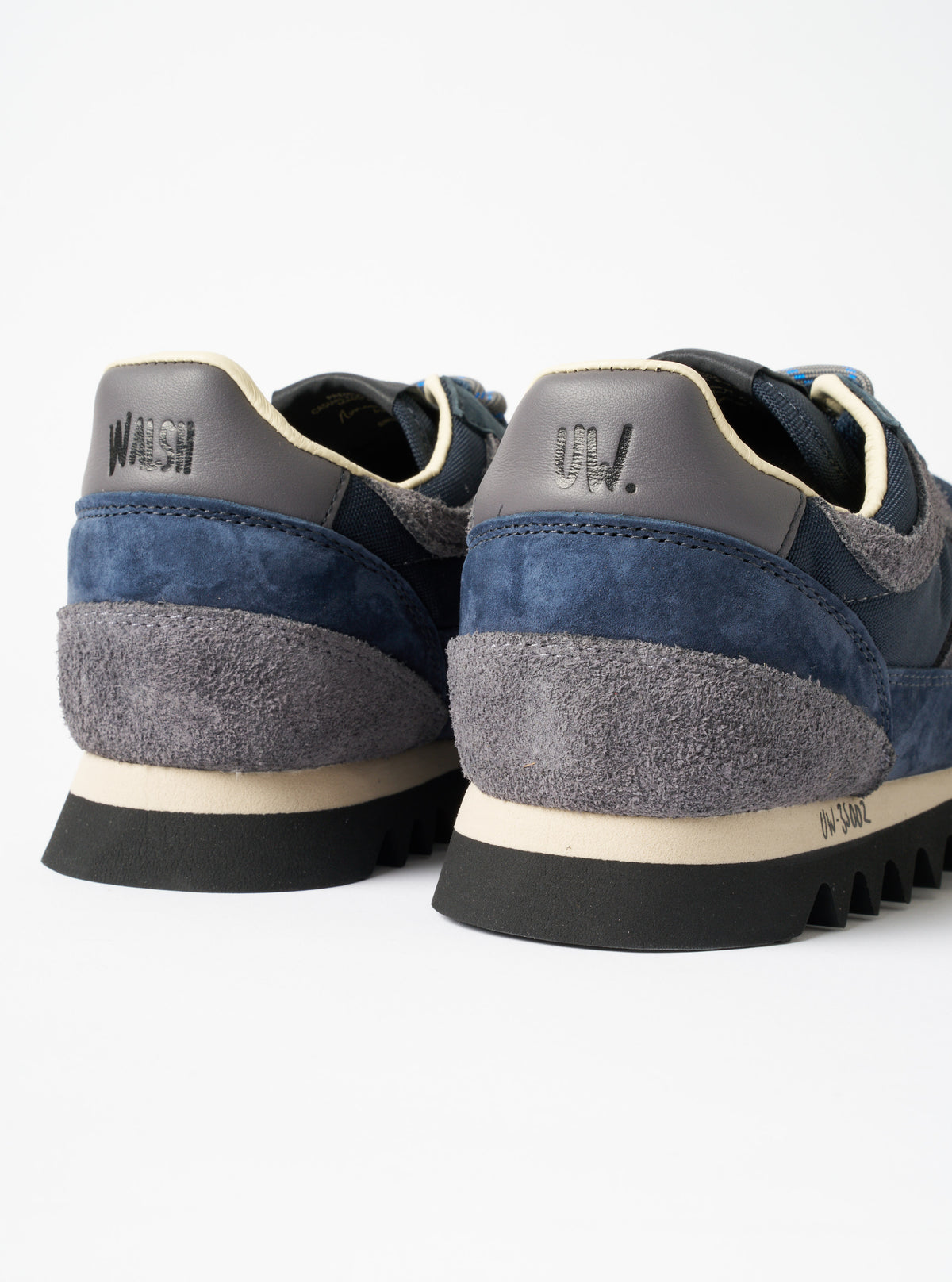 A full-length shot of a model wearing the Walsh x Universal Works Ensign in Navy Leather/Canvas sneakers in a minimalist studio setting, demonstrating how the navy footwear pairs with contemporary casual trousers.