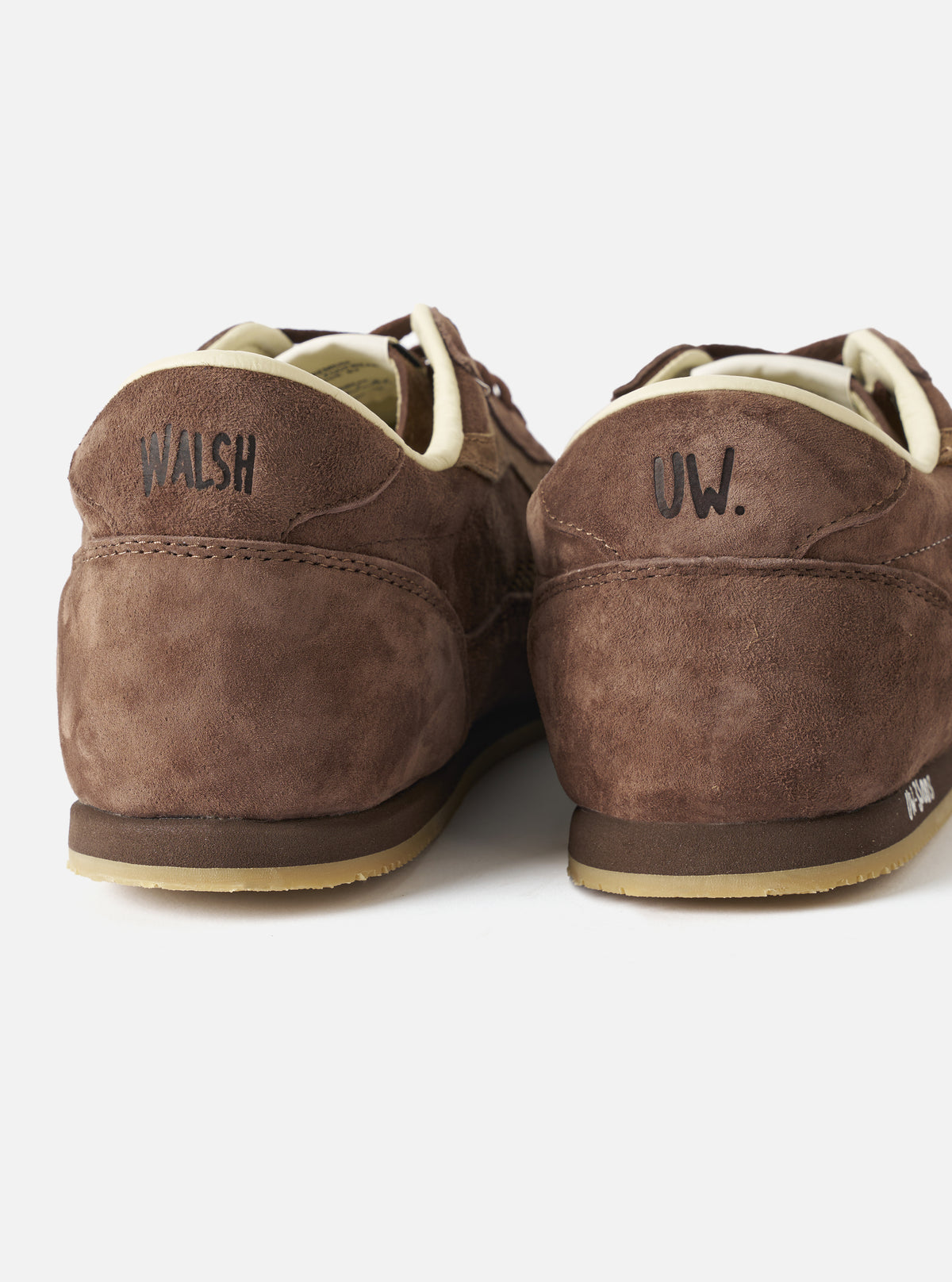 A macro close-up image showing the fine texture of the brown leather upper and co-branded details on the Walsh x Universal Works Cob in Brown Leather trainer against a clean white backdrop.