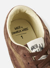 A full studio photograph of a model wearing the Walsh x Universal Works Cob in Brown Leather trainers standing against a plain light backdrop, highlighting the low-profile footwear styled with casual menswear pants.