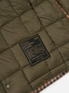 Close-up of the TAION x Universal Works Reversible Dark Olive Military Line Liner Jacket, showing the quilted dark olive lining and a branded tag. Houndstooth pattern visible on the edge.