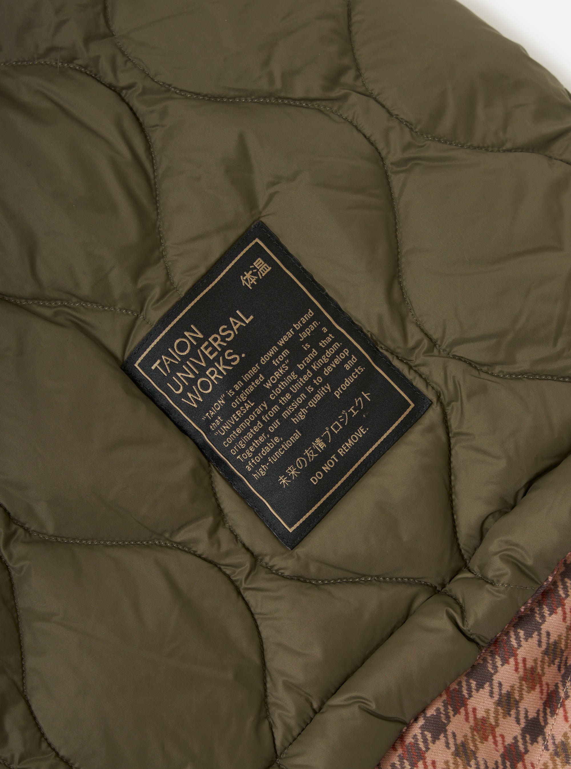 Close-up of the TAION x Universal Works Reversible Dark Olive Military Line Parka, showing the dark olive quilted lining and brand label. Part of the brown patterned outer layer is also visible.