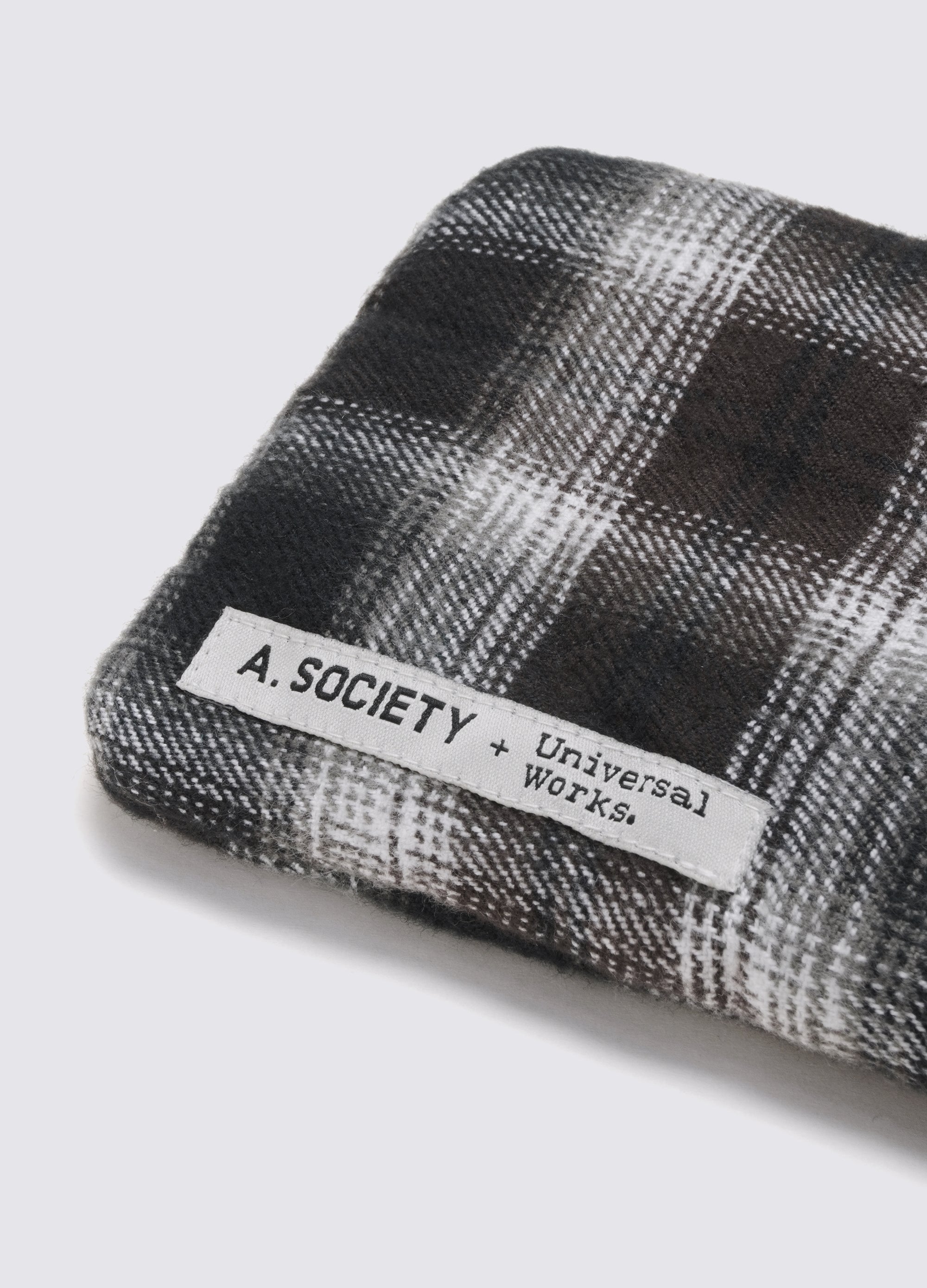 Close-up of A. SOCIETY x Universal Works Grey Aberdeen Sunglasses case. Grey and white plaid fabric. Label with brand names. Shot on a white surface.